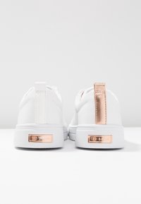 Baskets basses blanches avec languette de talon en or rose et étiquettes métalliques de la marque à l'arrière des semelles, vues de dos sur une surface blanche.