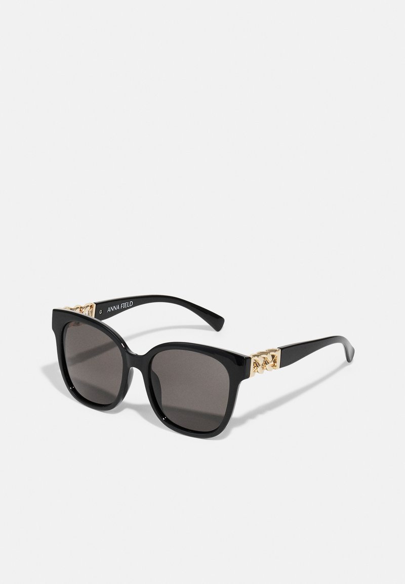 Black square sunglasses with gray lenses, featuring gold chain accents on the temples. Glossy finish, structured shape, and brand text on arm.