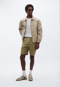 Μπεζ μπουφάν bomber πάνω από λευκό μπλουζάκι, χακί πράσινα σορτς και πολύχρωμα αθλητικά παπούτσια, στέκεται μπροστά σε απλό λευκό φόντο.