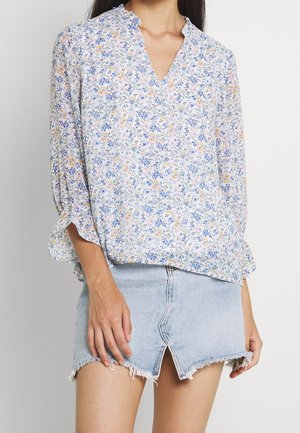 Femme portant un chemisier léger à manches longues à motif floral avec un col à volants et une mini-jupe en jean bleu clair déchirée avec une fente devant.