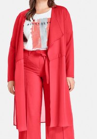 Mujer vistiendo pantalones anchos rojos y un cardigan largo rojo abierto sobre una camiseta blanca con el texto "#JUST BEY".