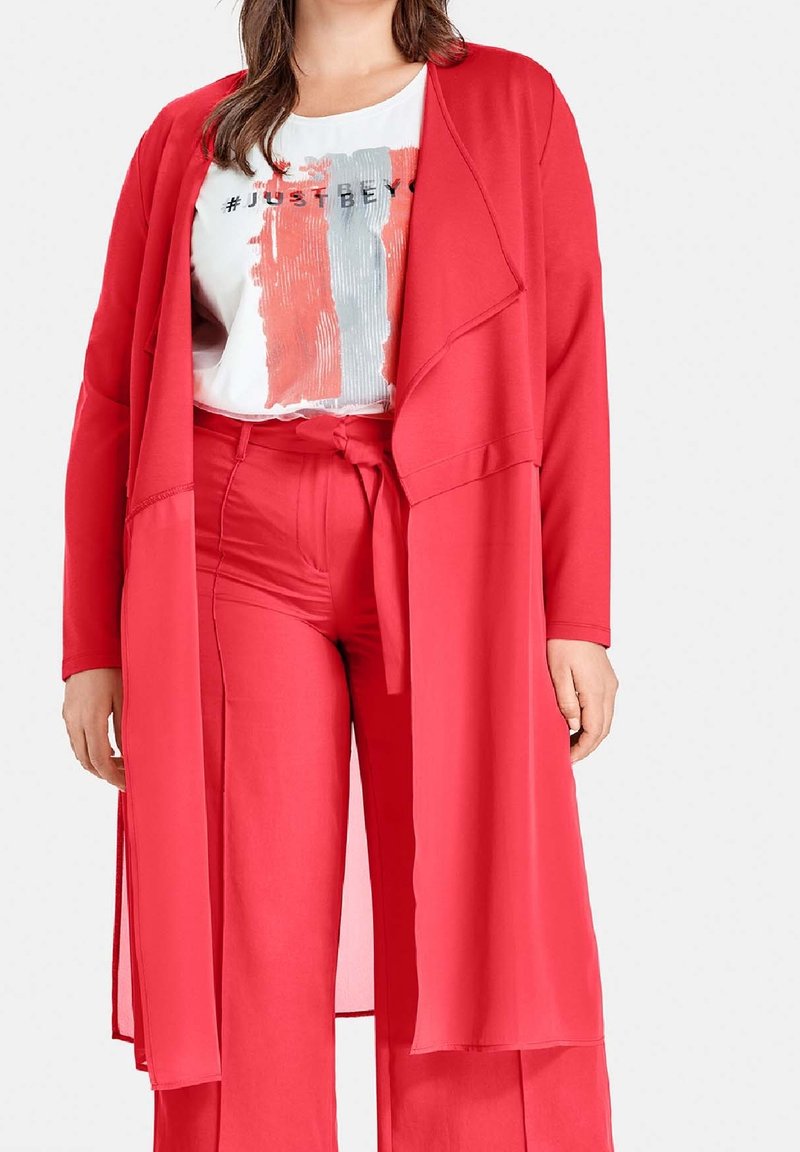 Mujer vistiendo pantalones anchos rojos y un cardigan largo rojo abierto sobre una camiseta blanca con el texto "#JUST BEY".