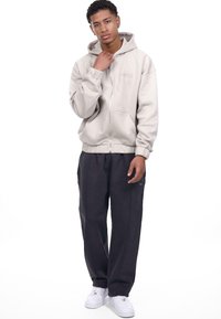 Beige ziptrøje med hætte og frontlommer, parret med mørke bukser med sideliste. Modellen har hvide sneakers på.