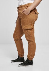 Brązowe spodnie cargo z elastycznym paskiem w talii, kieszeniami po bokach i ściągaczami przy kostkach, w zestawie z czarnymi sneakersami typu high-top. Gładka tekstura materiału.