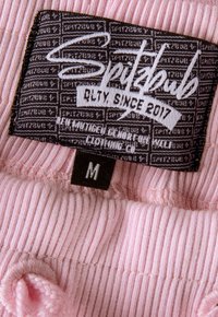 Das Etikett eines pinken gerippten Kleidungsstücks zeigt die Marke "Spitzbub" auf schwarzem Hintergrund mit weißem Text. Das Größenetikett liest "M" und befindet sich in der Nähe eines Strickdetails.