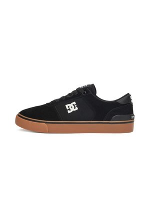 Zapatilla negra de caña baja con suela marrón, logo de DC en el lateral y cordones negros, vista desde el lado interior sobre un fondo blanco.