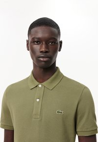 Olivgrön polo skjorta i texturerad bomull, med krage, tvåknappstängning och broderad krokodil-logotyp på vänster bröst.