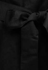 Μαύρο ύφασμα δεμένο σε ένα μαλακό, φαρδύ φιόγκο με ορατή υφή πλέξης και ελαφριές πτυχώσεις, που υποδηλώνουν λεπτομέρεια ρούχου.