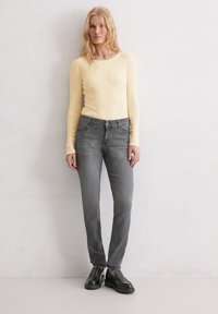 Femme blonde portant un haut à manches longues en rib courbé jaune pâle, un jean gris ajusté et des chaussures noires à enfiler, se tenant devant un mur blanc.