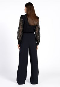 Vrouw met lang bruin haar, draagt een zwarte, wijde broek en een zwart top met doorzichtige, gebloemde lange mouwen, met haar rug naar de camera gekeerd tegen een witte achtergrond.
