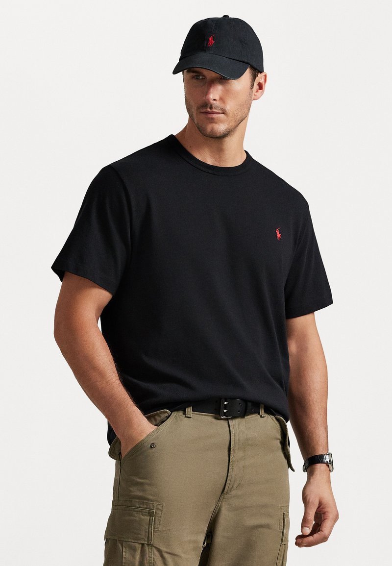 Μαύρη κοντομάνικη μπλούζα με μικρό κόκκινο κεντητό λογότυπο, συνδυασμένη με κηλιδωμένα παντελόνια καγκουρό και μαύρο καπέλο. Απλός, casual σχεδιασμός.