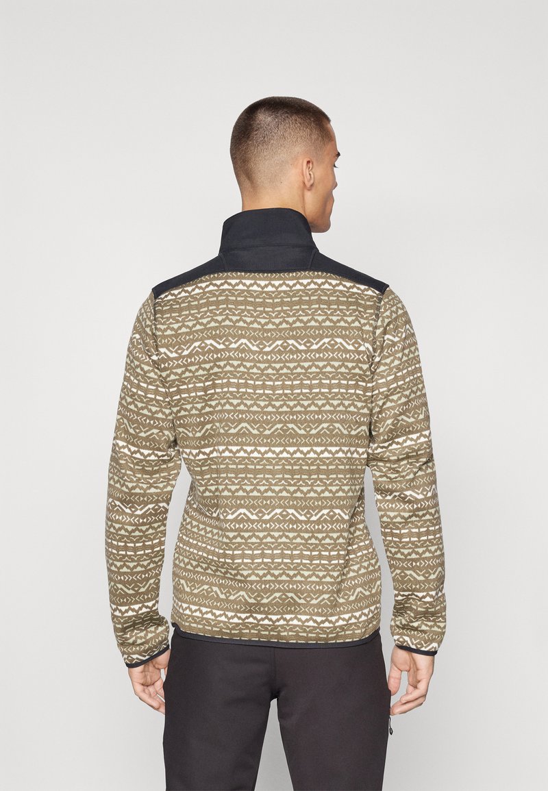 Hombre de espaldas, vistiendo una chaqueta a rayas marrón y blanca con cuello negro sólido y pantalones negros, contra un fondo claro y liso.