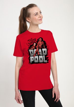 Ung kvinde iført rød Deadpool T-shirt med grafik og tekst, der siger "Ligner en plukket kalkun", stående mod en hvid baggrund.
