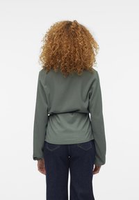 Haut à manches longues en maille côtelée de couleur vert atténué, avec une taille cintrée et des manches amples, associé à un jean bleu foncé. Fond uni.