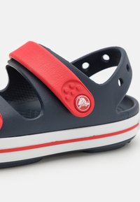 Sininen ja punainen kumitossu, jossa kaksi säädettävää hihnaa, kuvioitu pohjallinen, valkoinen välipohja ja pyöreä logokoriste hihnassa.