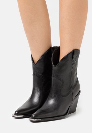 Sorte læderankelstøvler med spidse tæer, metalhætte på spidserne og kilehæle, båret på bare ben.