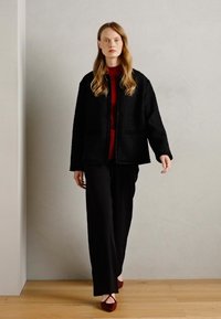 Veste noire texturée avec deux poches avant, portée sur un top rouge et un pantalon large noir. Le mannequin porte des chaussures rouges, debout sur un sol en bois.