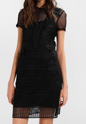 Woman wearing a black knee-length dress with floral lace appliqué and sheer short sleeves, standing against a plain light background.