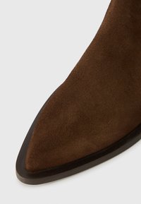 Barna velúr bokacsizma hegyes orral és sima, sötét gumitalppal. A textúrája puha, a dizájn letisztult, minimális díszítésekkel.