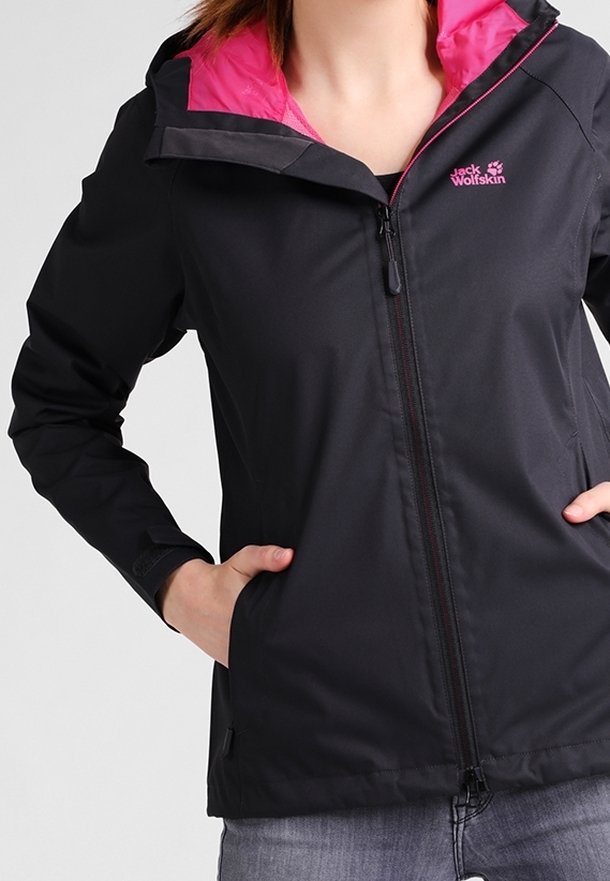 Veste noire avec doublure intérieure rose portée par une personne les mains dans les poches, montrant la fermeture éclair à l'avant et le logo Jack Wolfskin sur la poitrine.