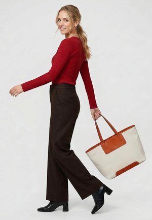 Žena v červeném topu s dlouhým rukávem a černých kalhotách kráčí a drží velkou béžovo-hnědou tašku s dvěma uchy.