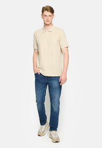 Beige polo tričko s krátkym rukávom, kombinované s modrou džínovou nohavicou a svetlými tenisovými topánkami; hladká textúra a klasický dizajn goliera.