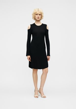 Γυναίκα με ξανθά σγουρά μαλλιά, που φοράει μαύρο φόρεμα με μακριά μανίκια και ακάλυπτους ώμους, καθώς και μπεζ ψηλοτάκουνα σανδάλια, στέκεται μπροστά σε λευκό φόντο.