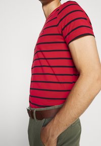 Rødstribet t-shirt med sorte vandrette striber, korte ærmer og rund halsudskæring. Båret med olivengrønne bukser og et brunt bælte.