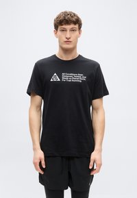 Μαύρο βαμβακερό μπλουζάκι με κοντά μανίκια, με λευκό τυπωμένο κείμενο και τριγωνικό λογότυπο στο μπροστινό μέρος. Κανονική εφαρμογή, σχέδιο με στρογγυλή λαιμόκοψη.