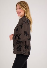Vrouw met lang blond haar draagt een donkergestreepte blouse met bloemenpatroon en zwarte broek, glimlachend en poserend in zijaanzicht tegen een effen achtergrond.
