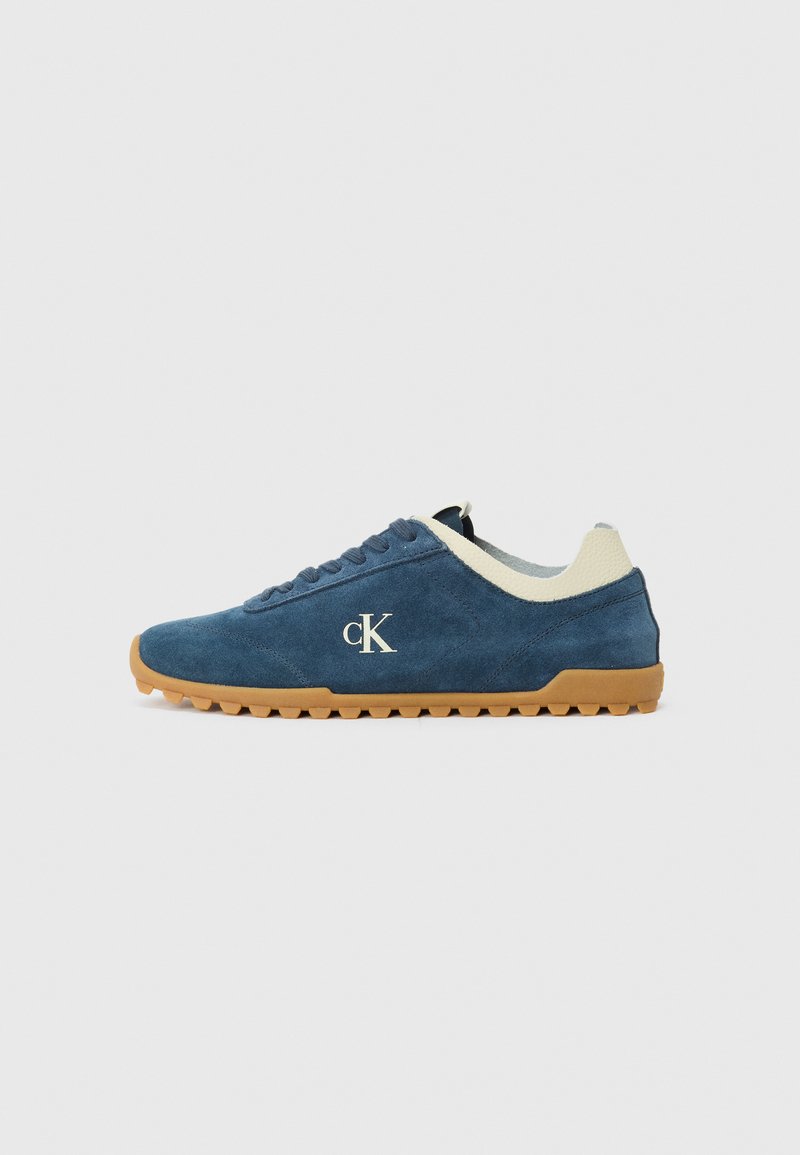 Blue suede sneaker with white tongue and heel accents, circular rubber sole with traction, and embroidered 'cK' logo on the side.