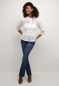 Blusa bianca in pizzo con maniche a tre quarti, chiusura frontale con bottoni, abbinata a jeans blu scuro e stivaletti marroni.