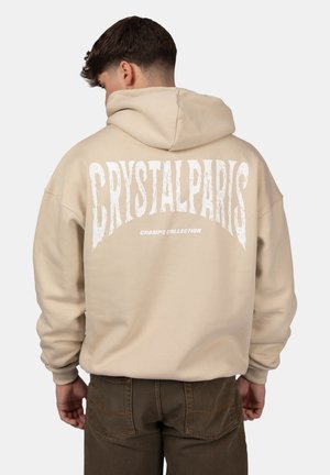 Felpa oversize beige con una grande grafica bianca che recita "CRYSTALPARIS" sul retro, dotata di cappuccio con lacci e polsini a costine.