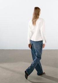White, long-sleeve shirt with side slits paired with light blue, loose-fitting jeans. Model wears black flats and has wavy hair.
