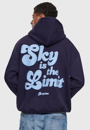 Mężczyzna noszący ciemnoniebieską bluzę z dużym jasnoniebieskim napisem na plecach "Sky is the Limit", w połączeniu z jasnoniebieskimi dżinsami.