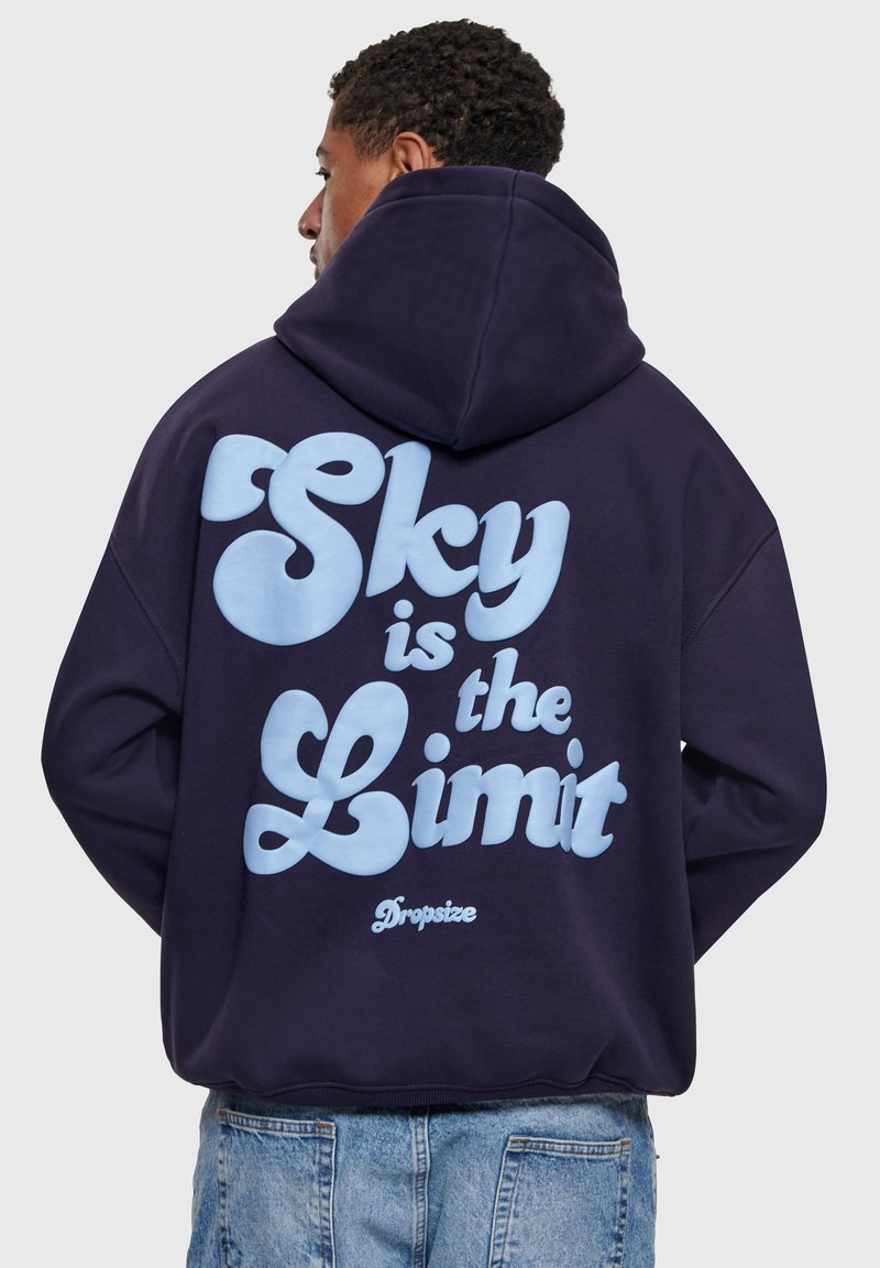 Homme portant un sweat à capuche bleu foncé avec un grand texte bleu clair au dos indiquant "Le ciel est la limite", associé à un jean bleu clair.
