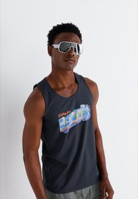 Svart tank topp med et fargerikt grafisk design som leser "Hilsen fra Levi's." Modell iført store hvite solbriller.