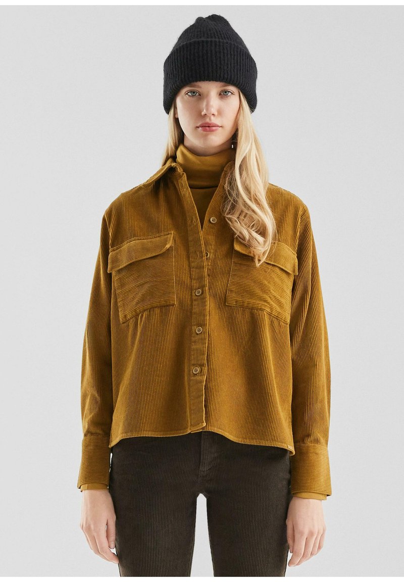 Vrouw in een mosterdkleurige corduroy knoopshirt over een bijpassende coltrui, zwarte beanie en donkere corduroy broek, staande tegen een effen achtergrond.