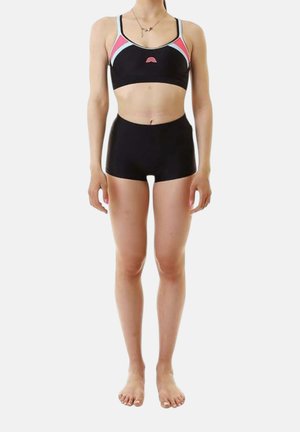 Donna in piedi a piedi nudi che indossa un reggiseno sportivo nero con dettagli rosa e azzurri e pantaloncini neri a vita alta su sfondo bianco.