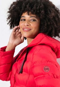 Röd pufferjacka med huva, kviltat mönster och svarta snoddar. Logopatch synlig på vänster ärm. Slät, glänsande tyg.