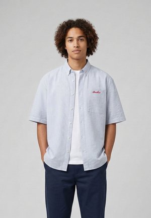 Junger Mann mit lockigem Haar steht mit den Händen in den Taschen, trägt ein hellblau gestreiftes, kurzärmeliges Hemd über einem weißen T-Shirt und dunkle Hose.