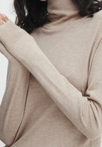 Maglione beige con collo a imbuto, maniche lunghe, polsini a coste e tessuto morbido e texturizzato. La vista ravvicinata mette in evidenza i dettagli della maglia.