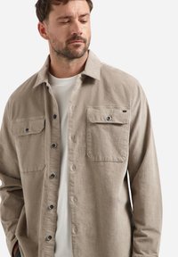 Man wearing beige buttoned shirt jacket over white crew neck shirt, looking down and to the side, hands in pockets.