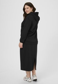 Femme aux cheveux bruns portant un hoodie noir et une jupe longue avec une fente à l'arrière, associée à des baskets blanches, se tenant contre un fond blanc.