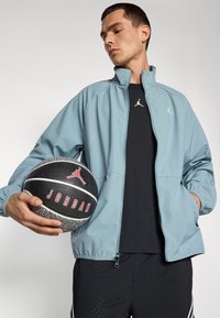 Chaqueta de chándal azul claro con cremallera y puños elásticos, camiseta negra con un logo y balón de baloncesto negro con acentos rojos y superficie texturada.