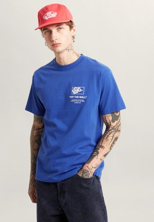 Blå bomulds T-shirt med korte ærmer, med et hvidt grafisk logo og tekst på brystet. Parret med mørke stofshorts.