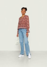 Blusa de mangas longas, com decote em V, na cor vermelho-escuro com padrão floral, combinada com jeans azul-claro de perna reta com fendas laterais. Bolsa branca e ténis.