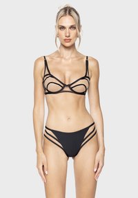 Vrouw met blond haar, gekleed in een zwarte mesh bh met bandjes en bijpassende slipje, staande tegen een effen lichte achtergrond, recht vooruit kijkend.
