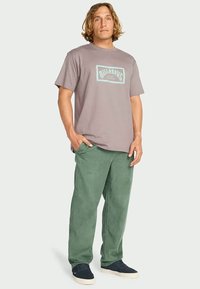 T-shirt cinza de manga curta com uma estampa azul na frente, combinado com calças de cortura verdes e ténis azuis-marinho. Conjunto simples e casual.