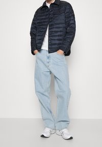 Mężczyzna w czarnej pikowanej kurtce, białej koszuli, jasnoniebieskich luźnych d�żinsach i białych sneakersach stoi z rękami w kieszeniach.